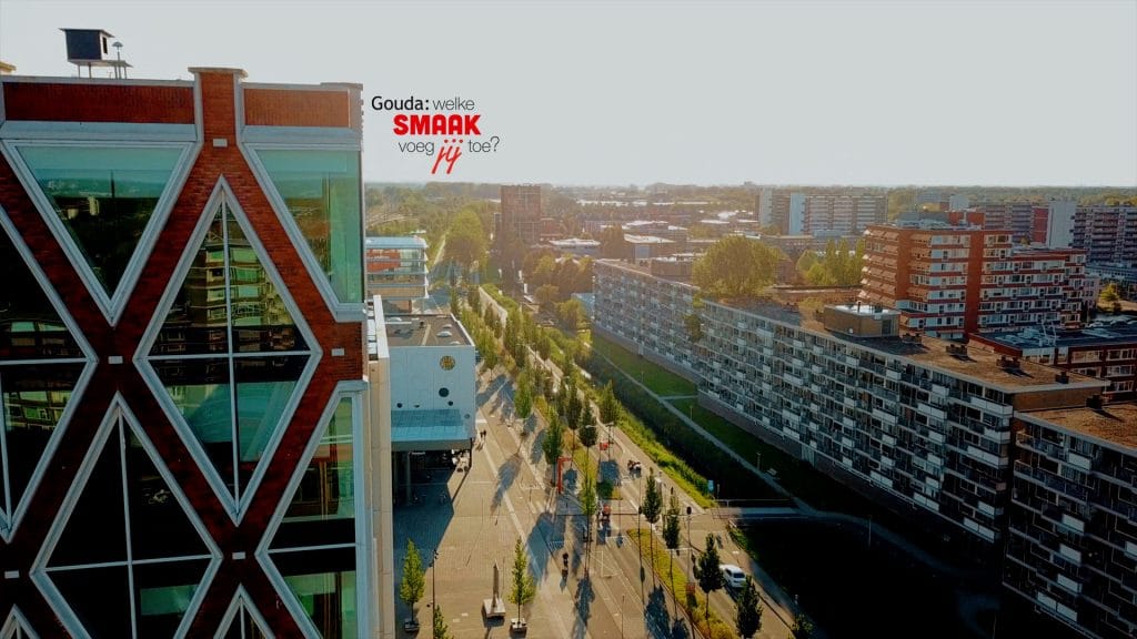Dronefoto van een gedeelte van het Huis van de Stad met op de achtergrond de bioscoop en rechts uitzicht over verschillende flats. In het midden het logo Gouda: welke smaak voeg jij toe?