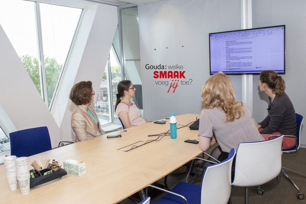 Vier collega's aan tafel in een vergaderkamer in het Huis van de Stad die naar een scherm aan de muur met een presentatie erop kijken. In het midden het logo Gouda: welke smaak voeg jij toe?