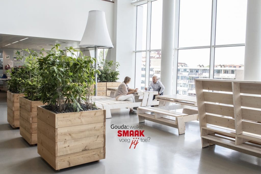 Twee collega's overleggen met elkaar zittend op picknickbanken in het Huis van de Stad, met rechts 3 plantenbakken. In het midden het logo Gouda: welke smaak voeg jij toe?