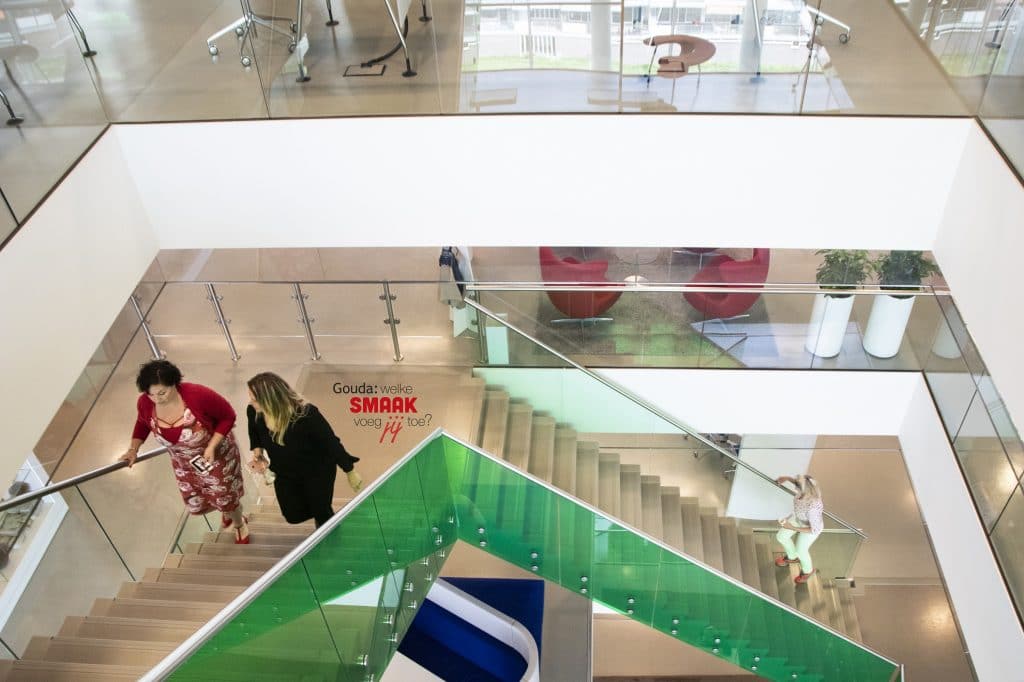 Het trappenhuis in het Huis va de Stad van bovenaf gezien met links 2 collega's die de trap op lopen. Daarnaast het logo Gouda: welke smaak voeg jij toe?