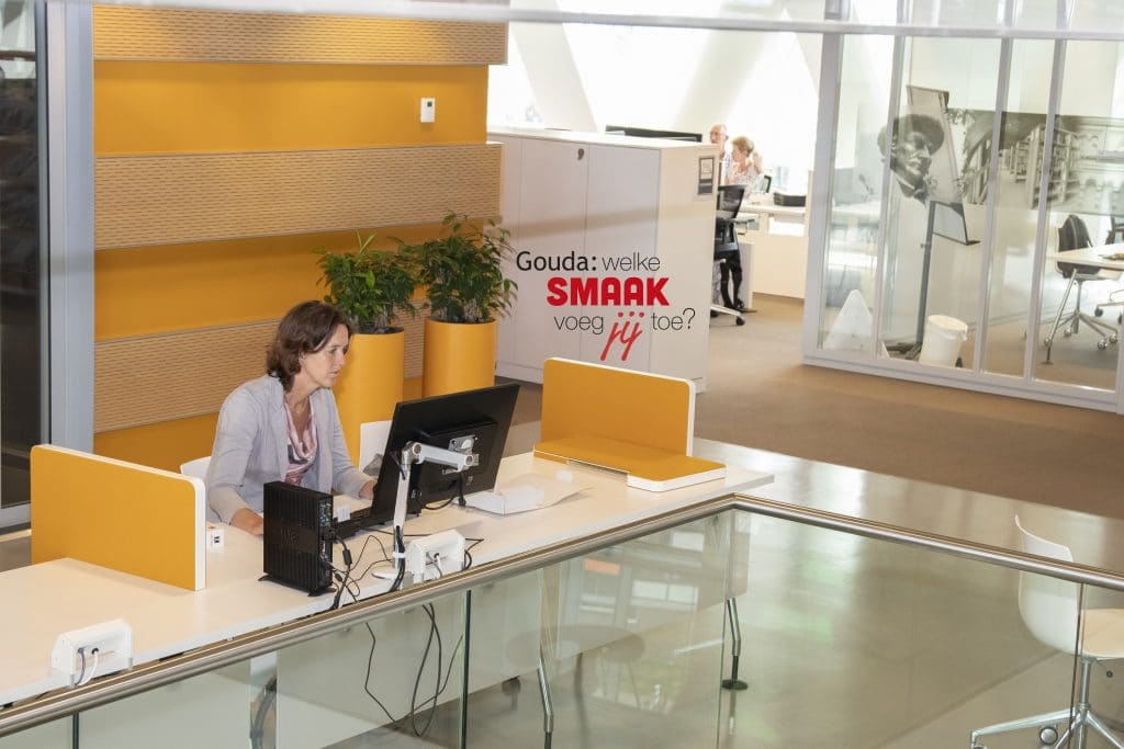 Een collega aan het werk achter een computer in het Huis van de Stad. In het midden het logo Gouda: welke smaak voeg jij toe?
